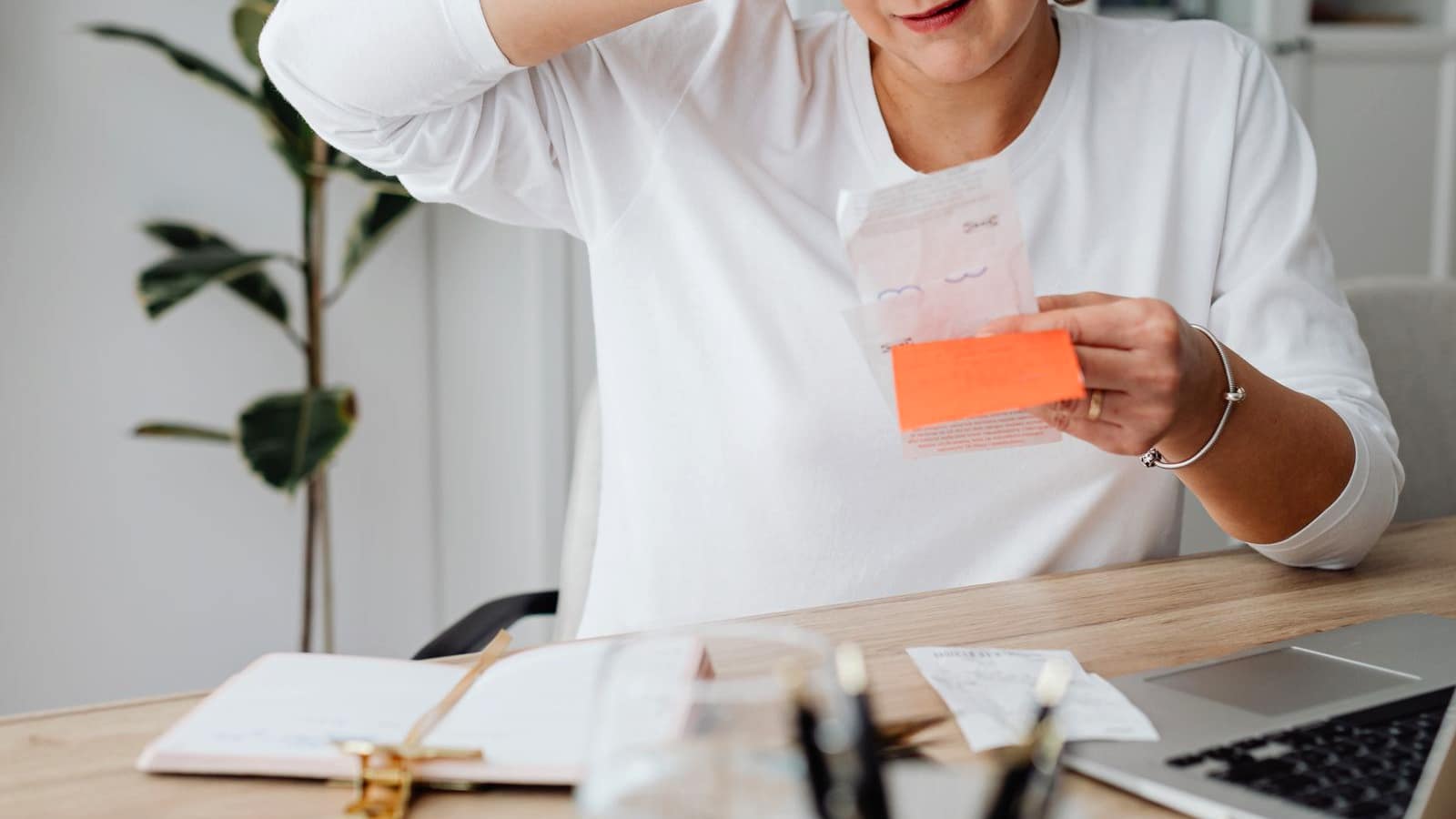 A person at a desk holding a receipt with one hand pressed to their head, surrounded by paperwork