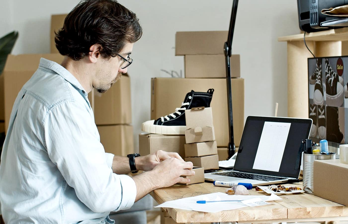 A person packing and photographing a product for an online store, surrounded by shipping boxes and a laptop