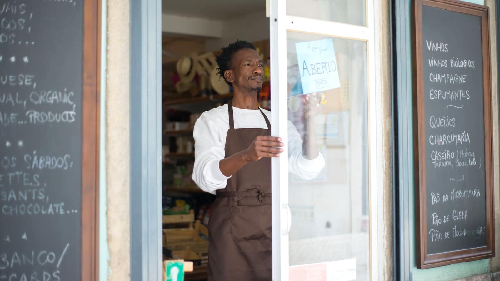 A shop owner in an apron putting up a sign in the doorway of a small food store