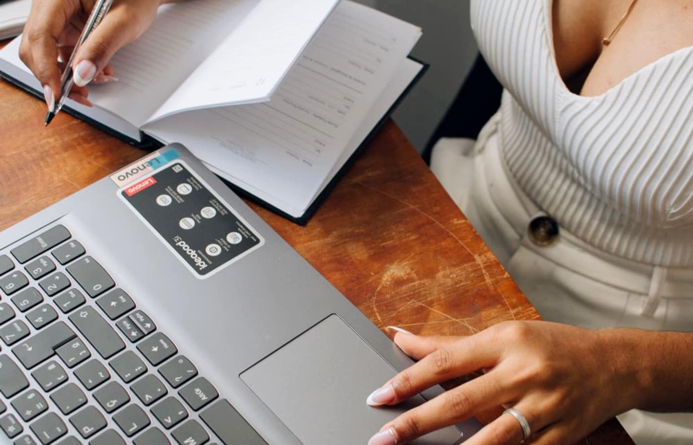 A person writing figures in a notebook beside a laptop on a wooden desk.
