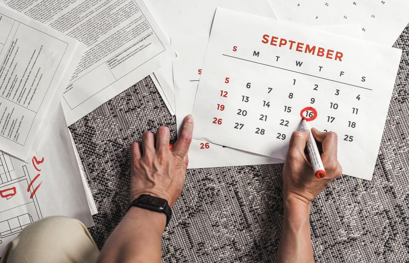 A person circling a date on a wall calendar with paperwork spread on the desk below