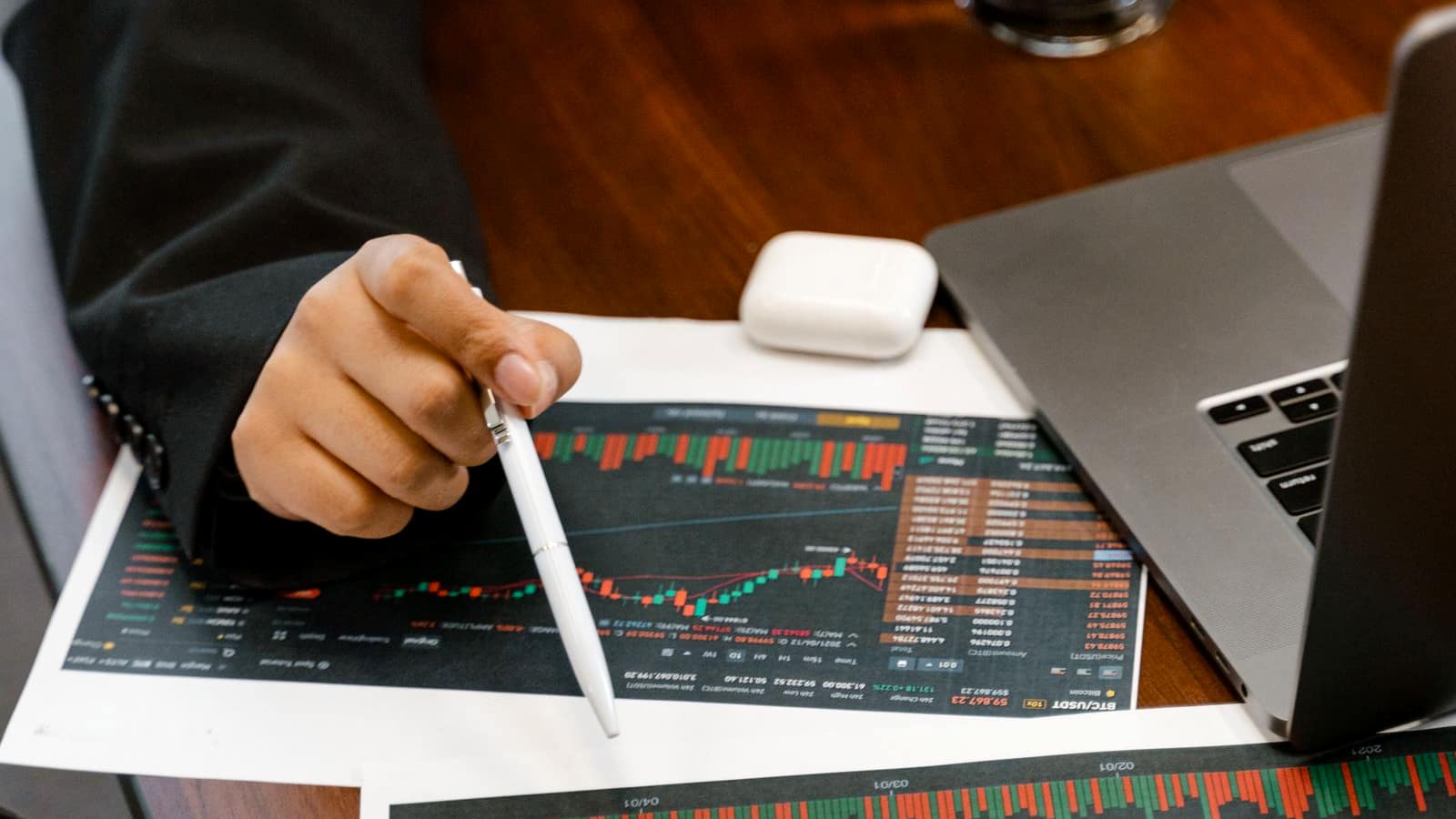 A person in a suit pointing a pen at printed financial charts next to a laptop
