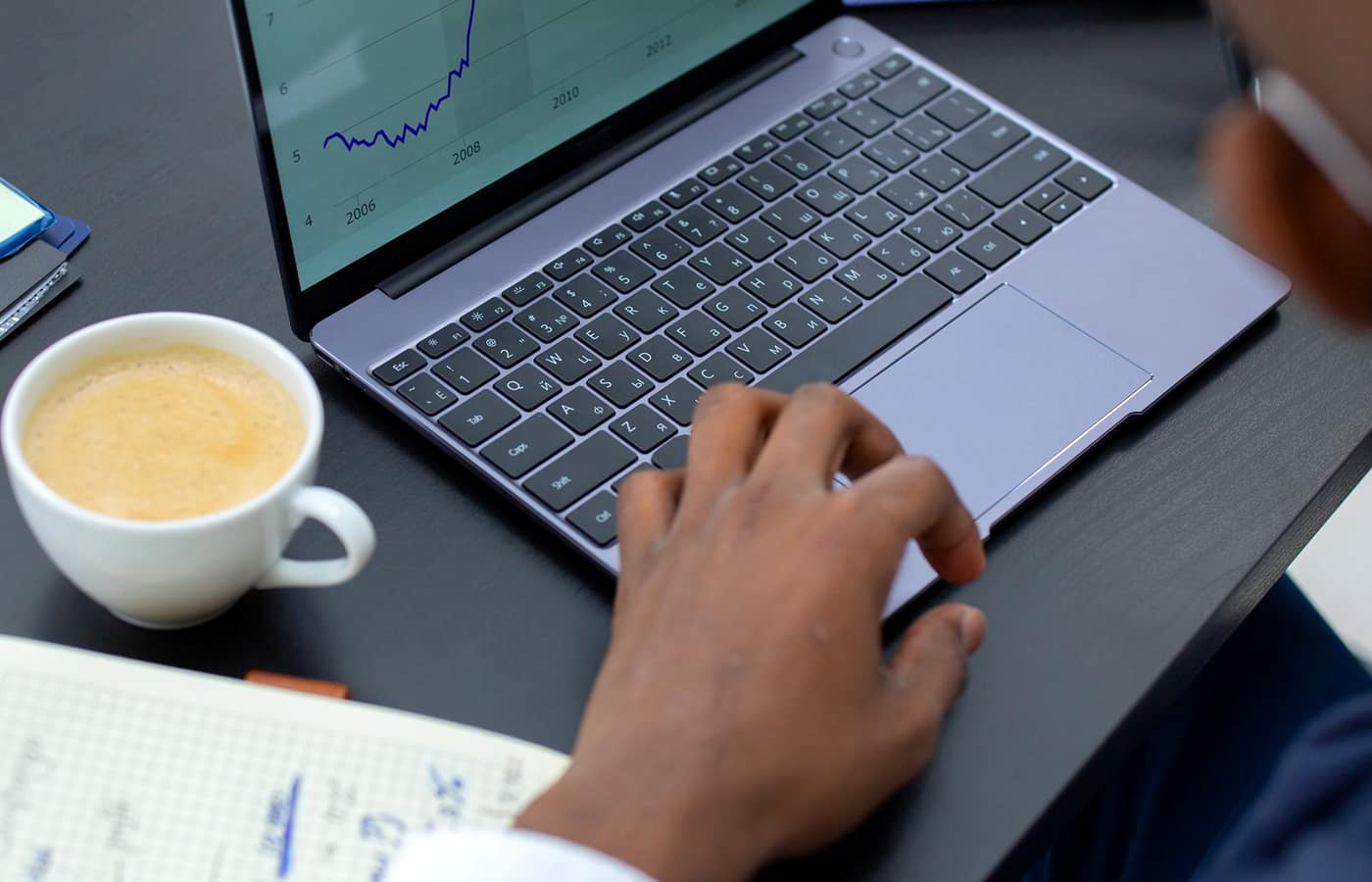 Hands working on a laptop showing a line chart, with a coffee cup and a notebook of figures nearby