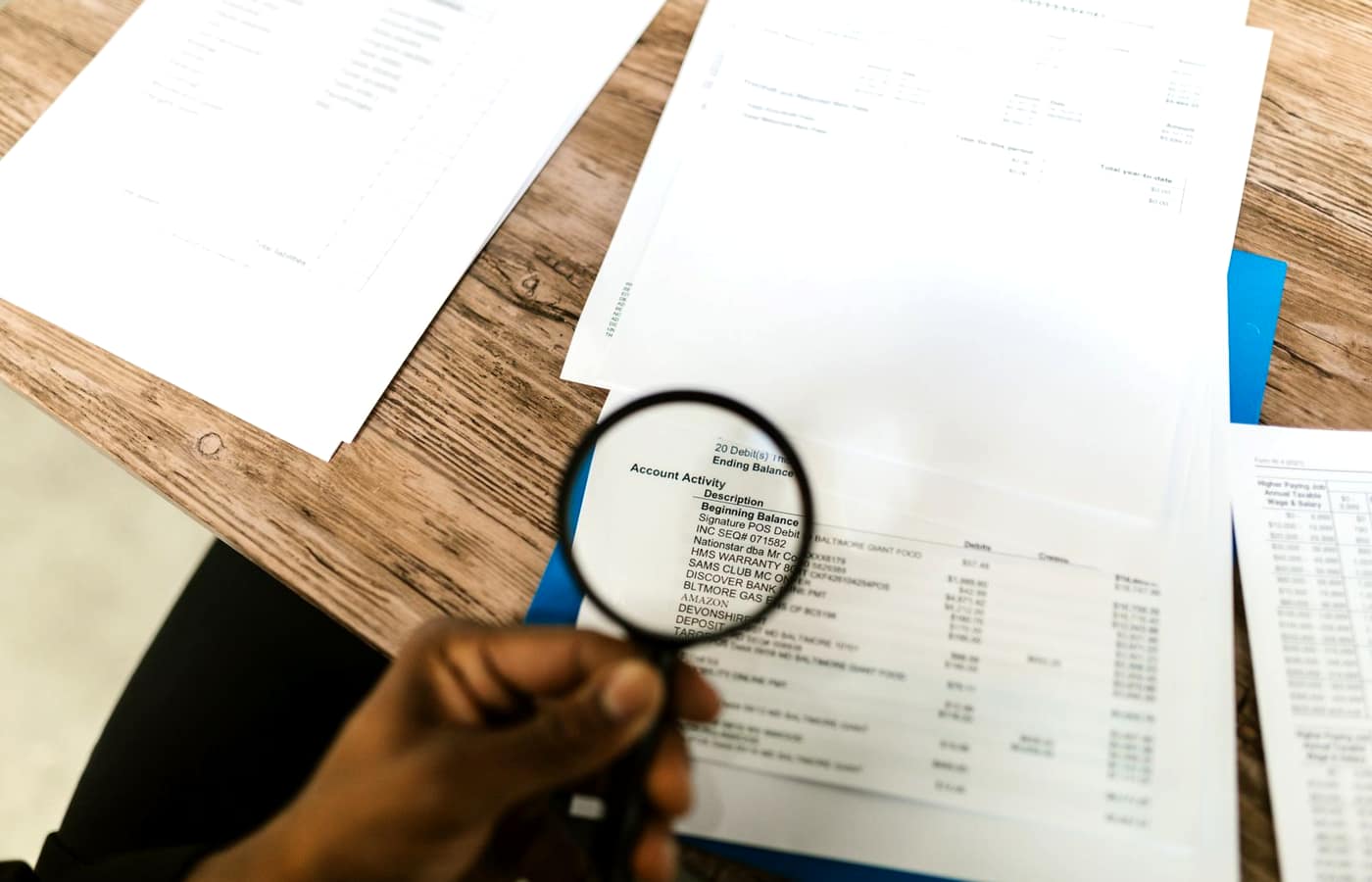 A hand holding a magnifying glass over a printed account-activity statement on a wooden desk
