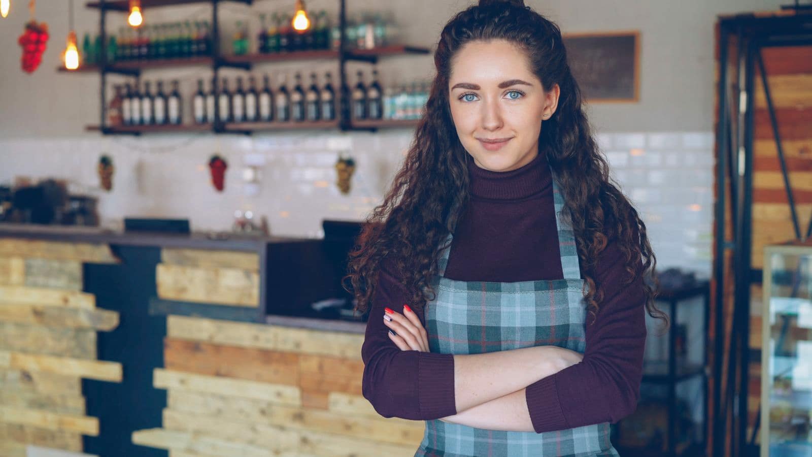 A small business owner in an apron standing with arms folded inside their cafe